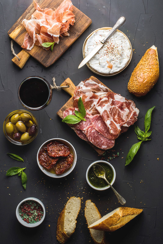 Wine snack set. Glass of red, meat selection, mediterranean olives, sun-dried tomatoes, baguette slices, camembert cheese and spices on black background, top view